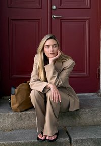 Beige räätälöity puku, jossa on leveät housut, istuu kivisillä portaille. Mustat flip-flopit ja ruskea käsilaukku. Taustalla punainen ovi. Sileä pinta.