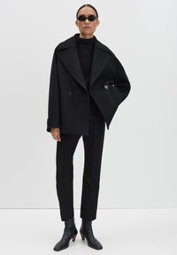 Cappotto nero doppiopetto con ampi rever, indossato su un maglione nero a coste, abbinato a pantaloni neri attillati e eleganti stivaletti neri alla caviglia.