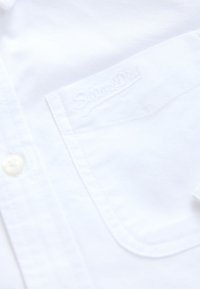 Gros plan d'une poche de chemise blanche à boutons avec un logo « Superdry » subtilement embossé au-dessus de la poche.