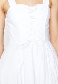 Robe blanche sans manches avec un lacet devant et un nœud à la taille portée par une personne avec un tatouage "be kind" sur le bras gauche.