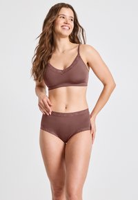 Donna sorridente con capelli lunghi, che indossa un reggiseno in pizzo mauve abbinato e mutandine a vita alta, in piedi su uno sfondo semplice.