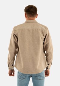 Man in een beige overhemd met lange mouwen en lichtblauwe jeans, staand met zijn rug naar de camera tegen een effen witte achtergrond.