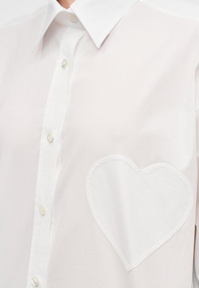 Λευκό πουκάμισο με κουμπιά και μυτερό γιακά, με τσέπη σε σχήμα καρδιάς στην αριστερή πλευρά. Κατασκευασμένο από ελαφρύ ύφασμα με λεία υφή.