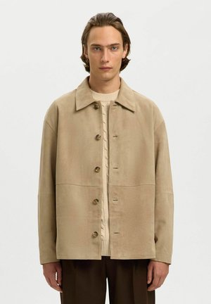 Jeune homme aux cheveux bruns mi-longs portant une veste beige boutonnée par-dessus un pull crème en maille torsadée et un pantalon foncé, faisant face à l'avant.