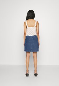 Žena s kudrnatými vlasy, oblečená v bílém topu bez rukávů s oranžovými puntíky a modré džínové sukni, stojící čelem ke bílé zdi.