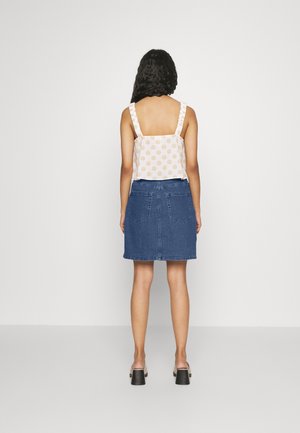 Mujer con cabello rizado, vistiendo una blusa sin mangas blanca con lunares naranjas y una falda de mezclilla azul, de pie frente a una pared blanca.