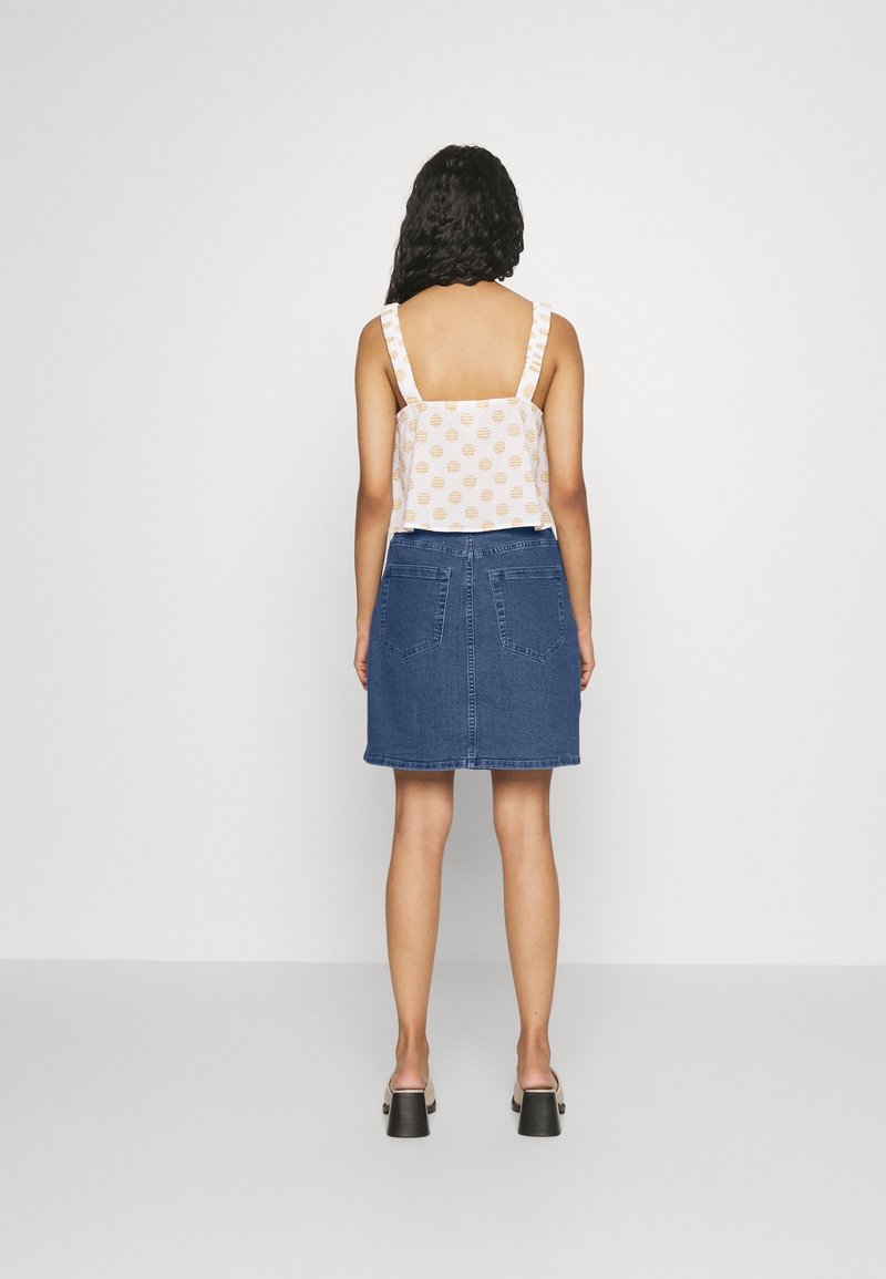 Žena s kudrnatými vlasy, oblečená v bílém topu bez rukávů s oranžovými puntíky a modré džínové sukni, stojící čelem ke bílé zdi.