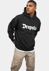 Sort hættejakke lavet af blødt stof med et hvidt broderet logo "Dropsia." Kængurulomme og ribbede manchetter. Båret med beige cargo bukser.