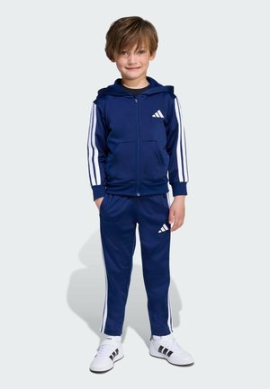 Chándal azul marino con rayas blancas en las mangas y los pantalones, que incluye una sudadera con cremallera y una cinturilla elástica. Incluye un par de zapatillas deportivas blancas.