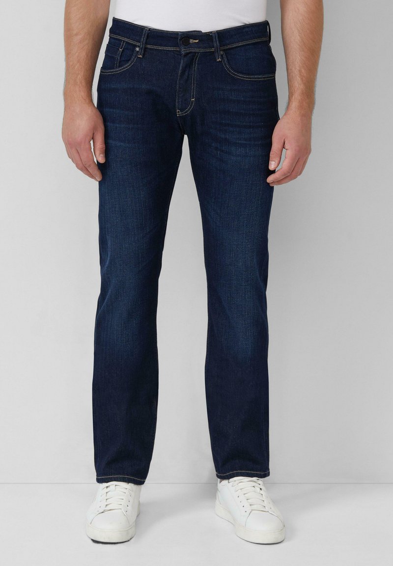 Dunkelblaue Denim-Jeans mit geradem Schnitt, fünf Taschen und kontrastierenden gelben Nähten. Kombiniert mit weißen Turnschuhen.