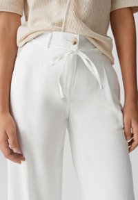 White linen trousers with a relaxed fit, featuring a drawstring waist, button closure, and front pockets. Smooth texture, minimal detailing.
