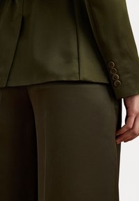 Olivgrön skräddarsydd kavaj med en slät yta, med en ficka och tre knappar på ärmarna. Matchas med byxor i samma färg.