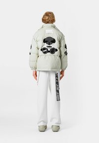 Chaqueta acolchada de color gris claro con detalles bordados en negro en la espalda y las mangas. Combinada con pantalones blancos que tienen una etiqueta lateral.