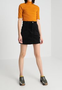 Maglione a collo alto arancione a maniche corte abbinato a una gonna in velluto a coste nera. Indossato con scarpe da ginnastica grigie con accenti gialli e suole marroni.