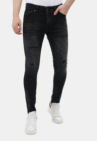 Zwarte skinny jeans met versleten plekken, subtiele gespikkelde details, vijf zakken en een knoopsluiting. Draagt met witte sneakers.