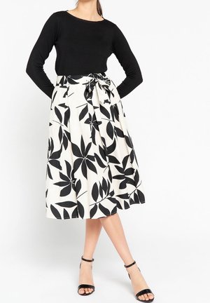 Mujer con top negro de manga larga, falda midi blanca con grandes estampados de hojas negras, atada a la cintura, y tacones negros con tira al tobillo.