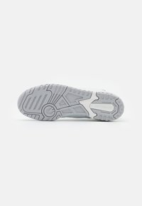 Semelle de basket New Balance grise et blanche avec des crampons à motifs et le nom de la marque visible sur un fond blanc uni.