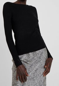 Haut noir à manches longues côtelées avec un col bateau, orné de boutons sur les poignets, associé à une jupe à motif noir et blanc.