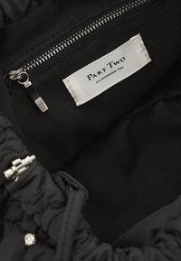 Must mehhitud must vooder kotis, millel on hõbedane lukuga tasku ja valge kanga silt, millel on kirjas "Part Two". Tekstuuriga, mati välimus.