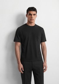 Jeune homme debout contre un mur pâle, portant un T-shirt noir uni et un pantalon noir, regardant droit devant avec une expression neutre.