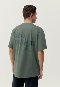 Zelené oversized tričko vyrobené z priedušnej látky, na chrbte s veľkým grafickým motívom "eko dizajn" a tmavým potlačou.