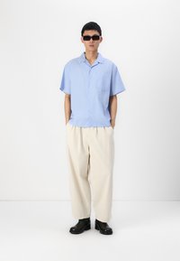 Chemise à manches courtes rayée bleu clair associée à un pantalon large crème et des bottines en cuir noires. Des lunettes de soleil complètent le look.