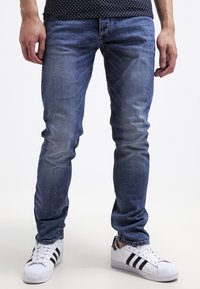 Slim-fit blå denimjeans med ljusblekningar och kontrasterande sömmar, i kombination med vita sneakers med svarta ränder.
