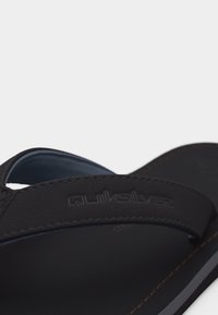 Črni Quiksilver natikač s prišitim trakom, na katerem je odtisnjen Quiksilver logotip, na belem ozadju.