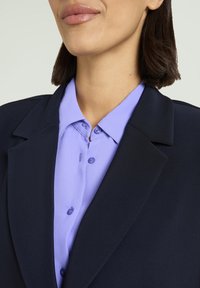 Marineblazer über einem hellvioletten Hemd mit Knopfdetails. Der Blazer verfügt über breite Revers und eine taillierte Passform. Glatte Stoffstruktur.