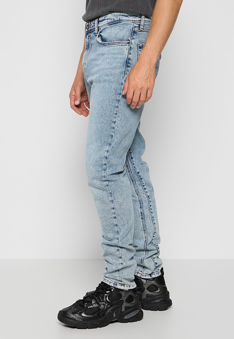 Seitenansicht einer Person, die leichte blaue Slim-Fit-Jeans und schwarze Polo-Sneaker trägt und auf einem hellgrauen Boden vor einer weißen Wand steht.
