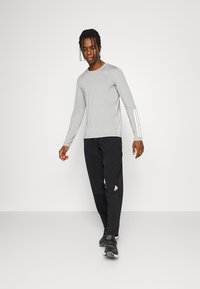 Camisa de manga larga gris claro con tres rayas blancas en una manga, combinada con pantalones de chándal negros que tienen un pequeño logo blanco. Zapatillas deportivas negras.