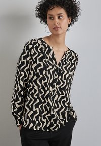 Schwarze und cremefarbene Bluse mit Knopfleiste, die ein Zickzackmuster aufweist, v-Ausschnitt und langen Ärmeln. Hergestellt aus leichtem Stoff mit lockerer Passform.