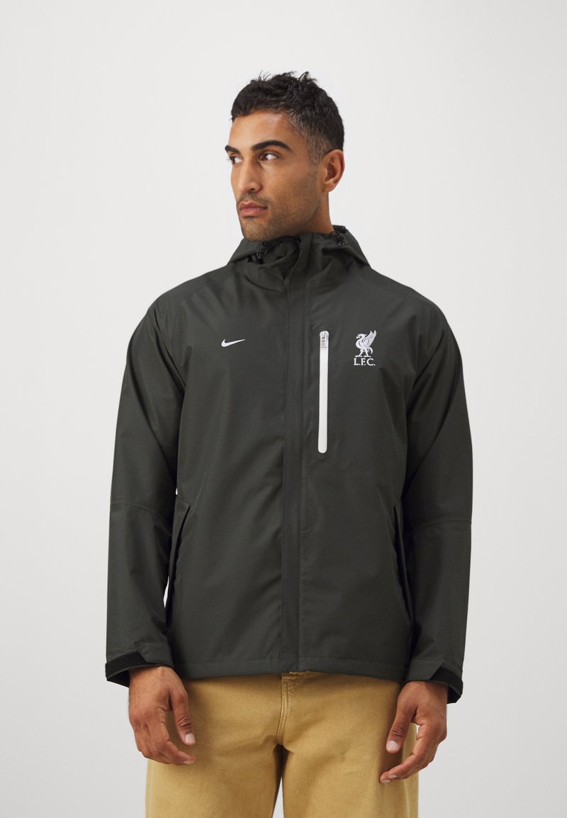 Schwarze wasserdichte Jacke mit Kapuze, weißem Frontreißverschluss, Liverpool FC-Logo auf der Brust und Nike-Logo auf der linken Seite. Glatte Textur.