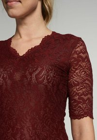 Burgunderfarbenes Spitzenoberteil mit V-Ausschnitt und kurzen Ärmeln. Filigranes Blumenmuster, gewellter Ausschnitt und leicht durchscheinende Textur.