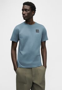 Μπλε κοντομάνικη μπλούζα από απαλό ύφασμα, με τετράγωνο μαύρο λογότυπο στο στήθος. Φορεμένη με χακί παντελόνι.