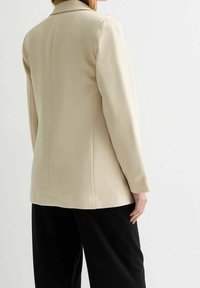 Blazer beige sur mesure avec une texture lisse, coupe ajustée, col rabattu et épaules structurées. Associé à un pantalon noir.