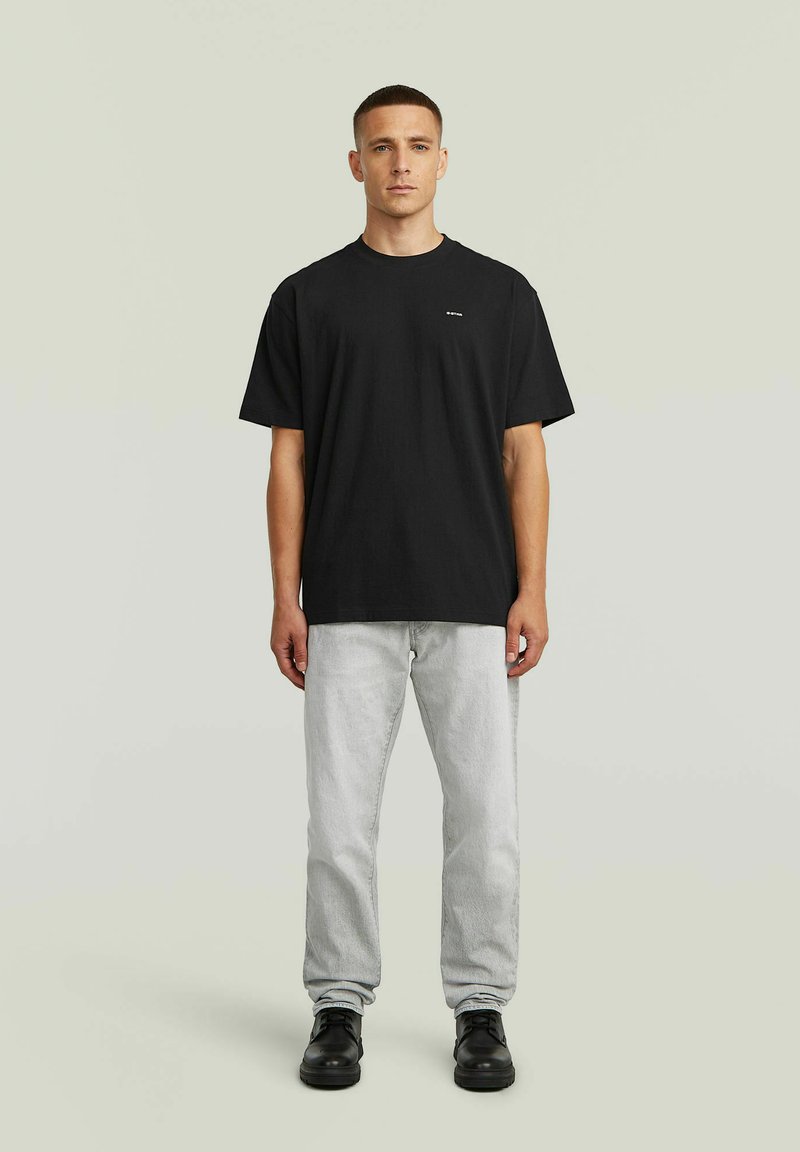 Uomo in piedi di fronte alla fotocamera, indossa una T-shirt nera a maniche corte, jeans grigio chiaro e scarpe nere su uno sfondo semplice.