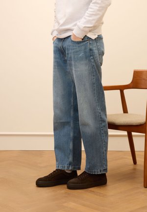 Light blue wide-leg jeans made of denim, featuring a relaxed fit, front pockets, and minimal fading, paired with black suede sneakers.