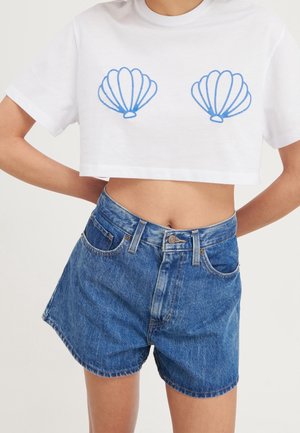 T-shirt blanche raccourcie avec un motif de coquillage bleu sur la poitrine, associée à un short en denim bleu taille haute avec un design classique à cinq poches.