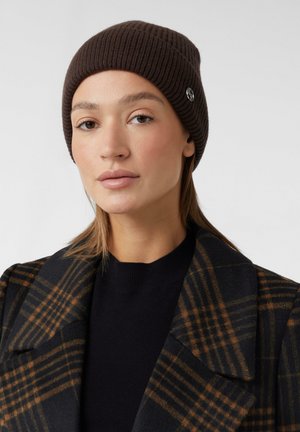 Brown ribbed beanie with a silver logo detail. Model wears a black sweater and plaid coat in shades of brown and black. Smooth texture.