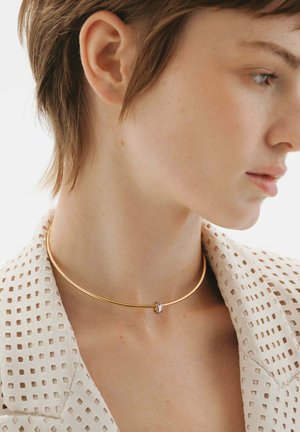 Collar de metal dorado con un acabado suave, que presenta un pequeño acento circular con un diseño contrastante, llevado en un cuello con cabello corto.