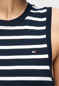 Μπλε ναυτικό αμάνικο τοπ με λευκές οριζόντιες ρίγες· διαθέτει ένα μικρό λογότυπο με κόκκινες, λευκές και μπλε πινελιές στο στήθος.