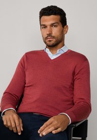 Czerwony sweter w serek wykonany z dzianiny, z długimi rękawami. Noszony na jasnoniebieskiej koszuli z kołnierzykiem, z widocznymi detalami mankietów.