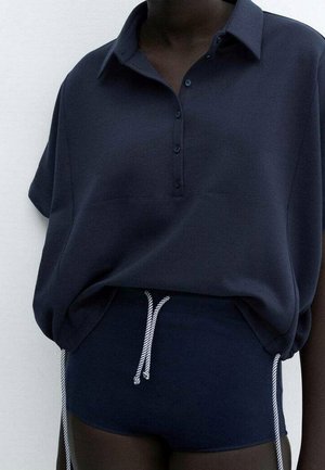 Mørkhudet person iført en løs mørkeblå skjorte med knapper og matchende shorts med hvide snore.