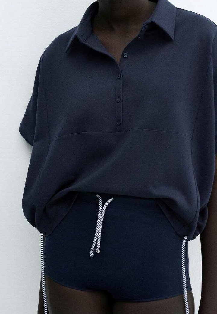Personne à la peau foncée portant une chemise ample bleu marine foncé avec des boutons et un short assorti avec des cordons blancs.