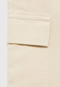 Beige tyg med diagonala strukturer, har en vikt ficka på höger sida och synlig sömnad längs sömmen.