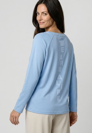 Frau lächelt über die Schulter, trägt ein hellblaues langärmliges Shirt mit vertikalem Text "WE ARE ALIFE" auf dem Rücken und beige Hosen.