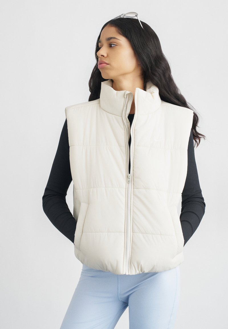 Λευκό αμάνικο μπουφάν με φουσκωτό ύφασμα, ψηλό γιακά, φερμουάρ μπροστά και πλαϊνές τσέπες, φορεμένο πάνω από μαύρη μπλούζα με μακριά μανίκια και ανοιχτό μπλε παντελόνι. Λείο υλικό.