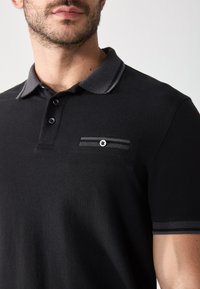 Schwarzes Poloshirt mit strukturiertem Kragen, zwei Knöpfen und einer Brusttasche mit Knopf. Akzente in Grau am Kragen und an der Tasche.