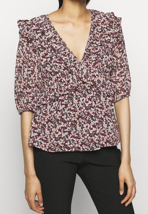Femme portant un chemisier cache-cœur à fleurs avec manches bouffantes et détails à volants, associé à un pantalon noir ajusté.